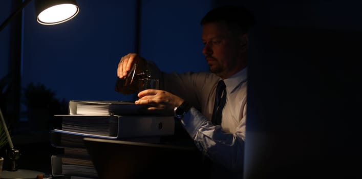 Portrait of tired businessman sitting indoors and pouring alcohol in glass. Man relaxing after intense workday with pile of paper folders full of corporation documents. Night at office concept