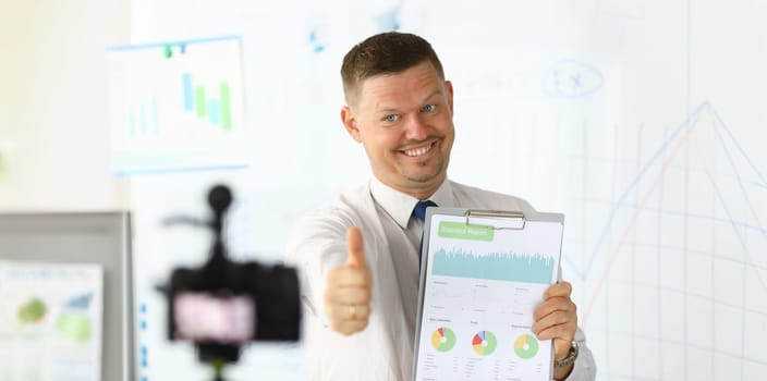 Portrait of cheerful man demonstrating statistics report with graphs, charts, diagrams and showing thumb-up for camera. Financial consultant making video for vlog. Blurred background