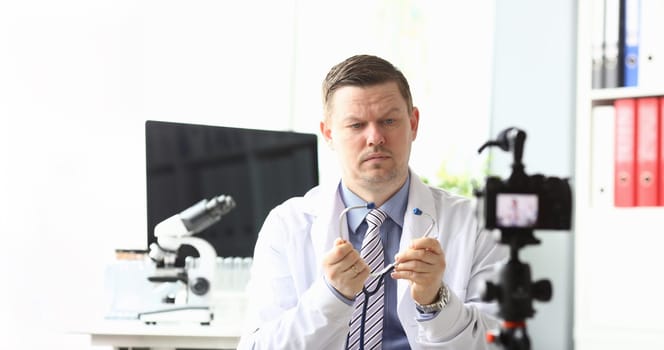 Portrait of sad hardworking doc blogger sitting in modern office and looking at stethoscope with question. Man in white robe wearing stethoscope. Hospital concept
