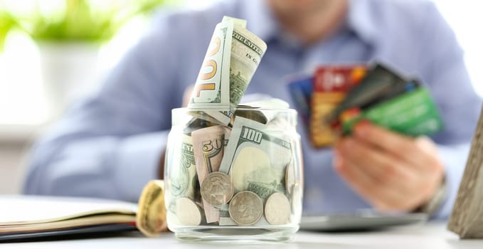 Big jar full of money stand at working table with male hand holding bunch of credit cards in background
