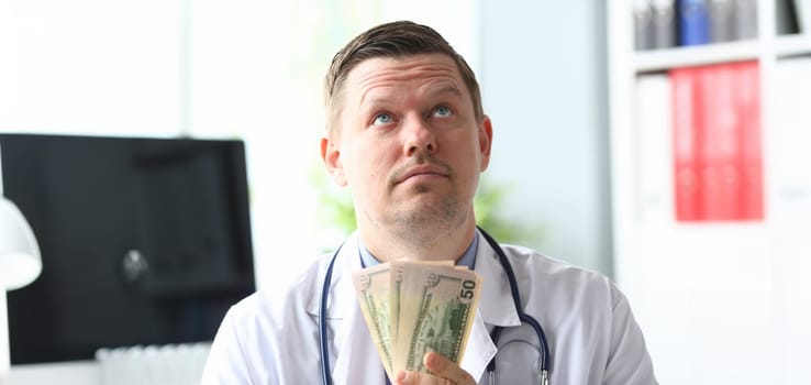Portrait of smart professional physician in modern hospital office holding fan of green banknotes and looking up with satisfaction. Healthcare and clinic concept