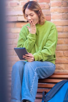 Vertical portrait beautiful Chinese teen student surprised using social media app on digital tablet outdoors. Girl sitting chatting on digital tablet