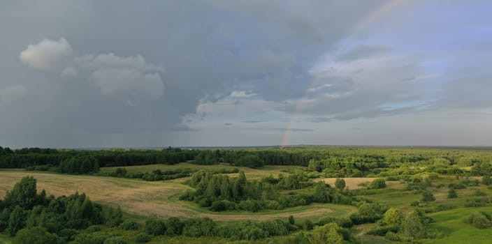Top view of meadow with road and green plants. Cloudy sky and beautiful rainbow above trees and bushes. Picturesque landscape scene of rain in summer time