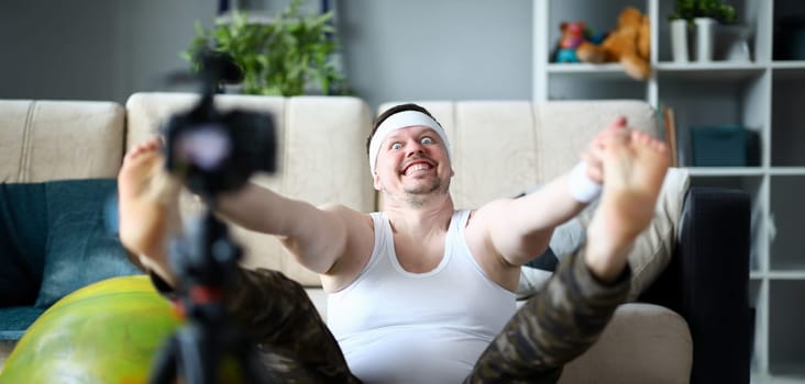 Vlogger freak shows his subscribers on camera how not to do the stretching correctly, holding his hands over his feet. Astonished face expression grimaces aganist home background concept