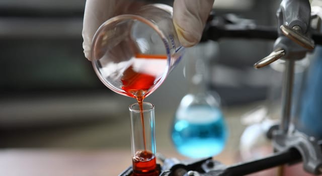 Focus on male hand in sterile gloves holding bulb. Pharmaceutist transfusing red fluid in glass flask. Medicine experiments and researches concept. Blurred background