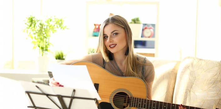 Millennial caucasian beautiful woman play guitar sit couch portrait. Misic scool concept home background.