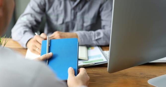 Male clerk holding blue notepad with silver pen talking to colleague close-up