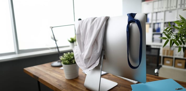Cabinet workplace with wooden table, documents and computer. White classic shirt and blue tie hanging on pc monitor. Things scattered at workspace. Difficult working day concept. Blurred background