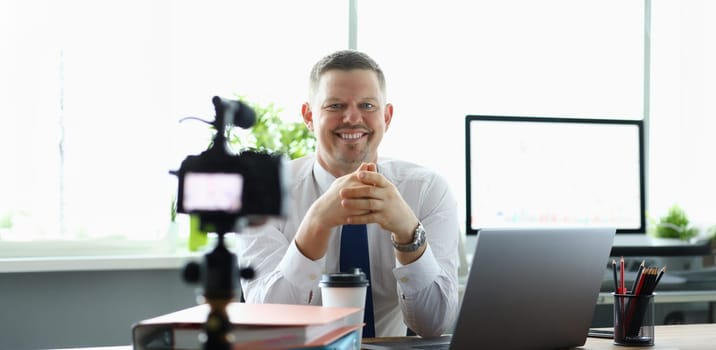 Portrait of cheerful man sitting at table with laptop and recording video for financial blog. Joyful guy giving online consultation for video blog. Copy space in computer screen. Blurred background