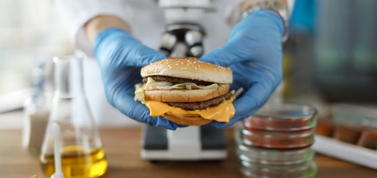 Male hand hold tasty burger in hand with blue protective gloves closeup. Testing food nutrition concept.