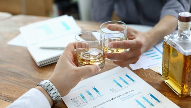 Focus on male hands holding glassful with whisky. Colleagues clinking glasses with alcoholic beverage. Business people celebrating signing profitable contract