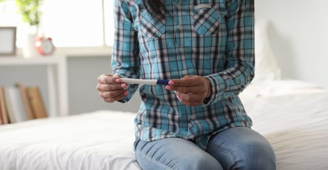 A lonely woman sits on bed and holds pregnancy test in her hand. Female maternity concept.