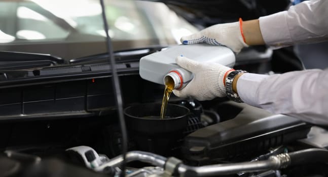 Focus on auto mechanic in gloves replacing and pouring fresh oil into engine at automobile service station. Vehicle inspection concept. Blurred background