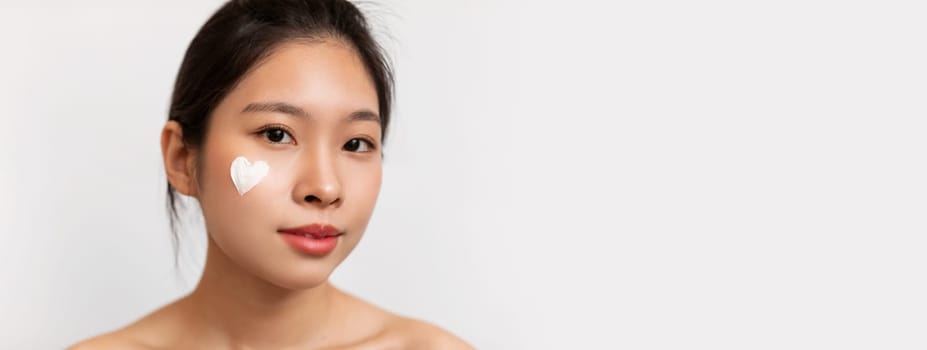Closeup portrait of pretty korean lady with moisturizer cream on her face in heart shape, woman testing new beauty product, white studio background. Face care concept, copy space