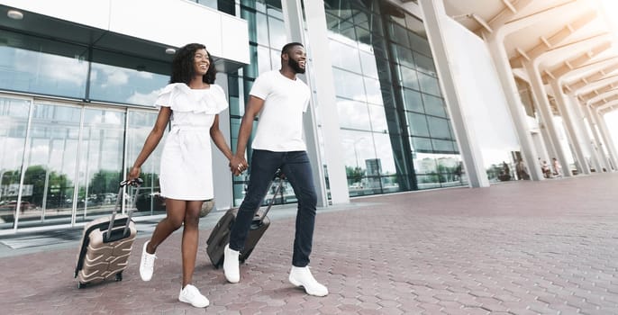 Black young couple smiling, walk towards the camera with their luggage in hand. They are outside of an airport building, with large glass windows and doors visible behind them, copy space