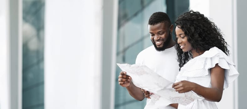 African American couple stands outside a building, looking at a map together. They are both smiling and appear to be happy. The woman is pointing at something on the map with her finger