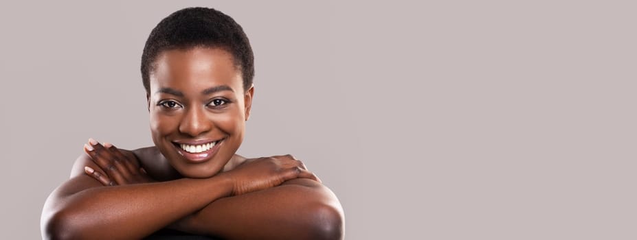 Beauty portrait. Charming nude black woman with flawless skin and white teeth resting her head on hands and looking at camera on grey background, copy space