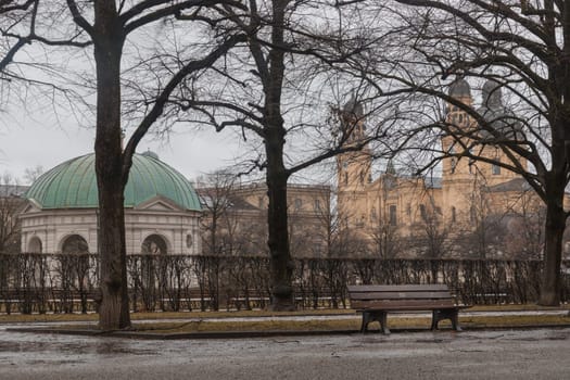 Munich, Germany - Dec 21, 2023 - Bench made wood and iron stand in the garden of the Munich Residenz with The Diana Temple (Dianatempel) and St.Cajetan and Adelaide in the background. Space for text, Selective focus.