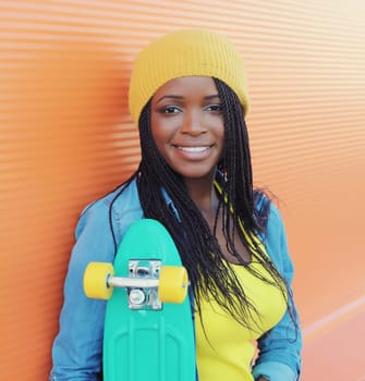 Summer portrait of stylish happy African young woman with skateboard, modern black teenage girl in yellow hat posing on orange background
