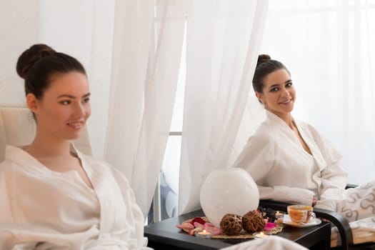 Two pretty young girls with nice bundles in white bathrobes sitting by the table laid for tea and relaxing shot in spa saloon