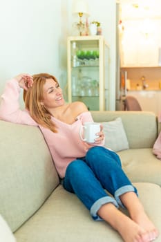 Vertical photo of a blonde mature adult happy relaxed woman drinking tea lying on the sofa