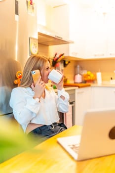 Vertical photo of a blonde caucasian female freelancer drinking coffee while working from home using mobile phone and laptop