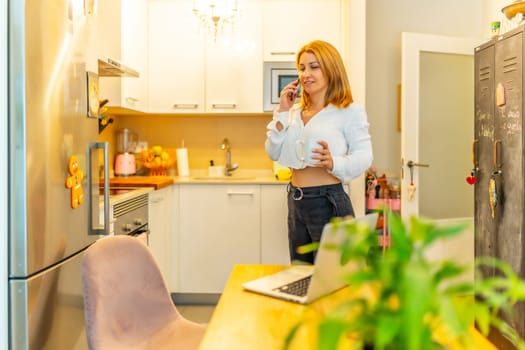 Blonde caucasian businesswoman talking to the mobile phone standing in a home office next to the kitchen