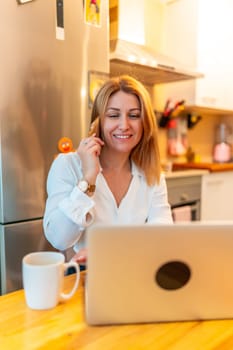 Vertical photo of a blonde caucasian adult smiling businesswoman taking to the phone while teleworking using laptop sitting in the kitchen
