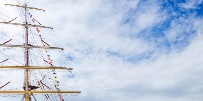 Sailing ship mast with flying nautical flags festival celebration decoration in a blue sunny sky and clouds, rigging of old tall ship with colorful bunting strung. Banner