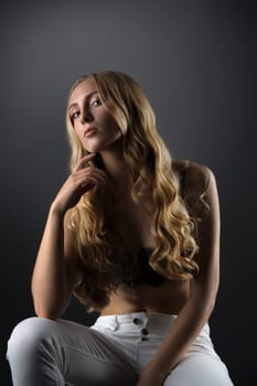 Beautiful blonde sitting on a cube dressed in white skinny jeans and bra studio portrait