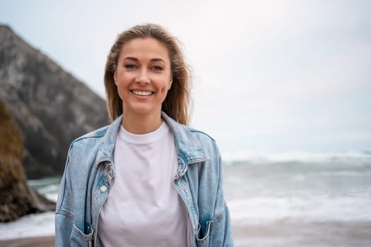 Smiling woman in denim shirt with sea and mountain in background. Portrait of young blond lady enjoying vacation summer vacation at beach. Happy girl looking at camera.