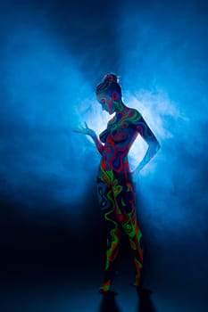 Slim girl posing in smoky studio her face and figure covered by bright fantastic ultraviolet color pattern bodyart.