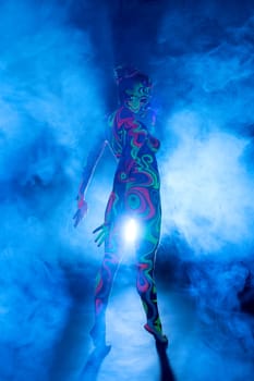 Slim girl posing in smoky studio her face and figure covered by bright fantastic ultraviolet color pattern bodyart.