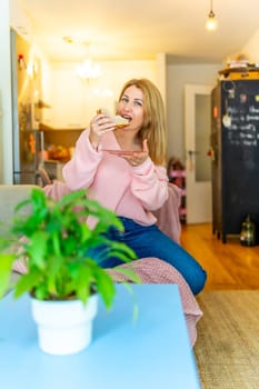 Vertical photo of a mature adult blonde beauty woman biting a healthy sandwich sitting on the sofa