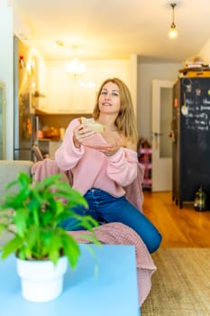 Vertical photo of a caucasian adult woman eating home made healthy sandwich sitting on the sofa at home