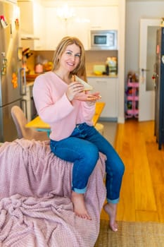 Vertical portrait of a mature adult caucasian blonde smiling comfortable woman eating healthy sandwich at home