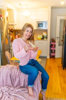 Vertical full length portrait of a blonde mature casual woman eating a healthy sandwich sitting on the sofa at home