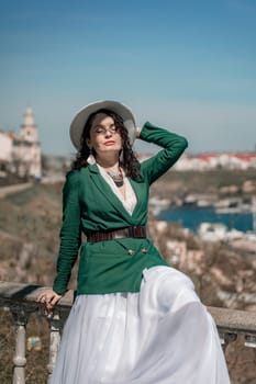 Woman walks around the city, lifestyle. A young beautiful woman in a green jacket, white skirt and hat is sitting on a white fence with balusters overlooking the sea bay and the city