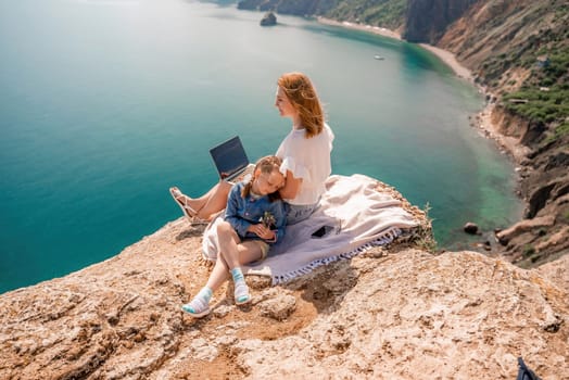 Freelance woman with her daughter working on a laptop by the sea, typing on the keyboard, enjoying the beautiful view, highlighting the idea of remote work