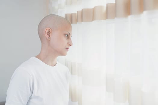 A young woman in her 30s with oncology who has lost her hair after chemotherapy stands by the window, copy space.