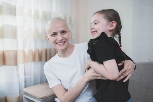 Young smiling bald woman with cancer spending time with her kid at home room. Oncology and family support concept.