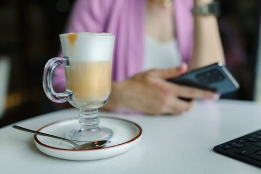 Business woman sitting in cafe with a laptop and smartphone, In the foreground, a tall glass with latte coffee close-up.