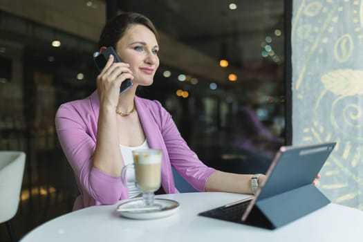 Attractive caucasian female freelancer talking on mobile phone while working in cafe behind laptop.