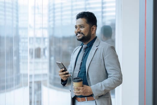 Indian businessman standing next to window using mobile phone wile coffee break.