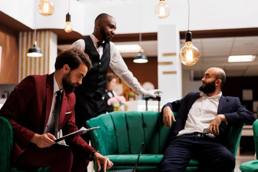 White collar worker reads files in lobby, working on official documents plan while he waits to see accommodation at hotel. Business representative reviewing speech notes, lounge area.