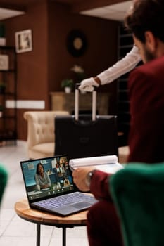 Businessman presents ideas on videocall, using online telework meeting in hotel lounge area reception. Corporate employee attends remote conference call, business trip internet chat.
