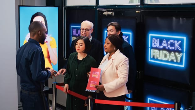 Anxious customers in line for deals on black friday, standing behind red tape at clothing store entrance. Impatient shoppers waiting for opening shop to start promotional shopping spree