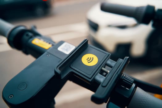 A close up of a phone holder on a scooter with a car in the background, resembling a small machine gun accessory on a larger automotive tire