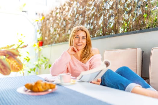 Adult caucasian blonde woman smiling at camera lying on a sofa in the outdoor yard enjoying breakfast and reading a book