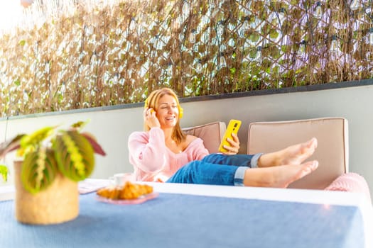 Caucasian adult woman lying on the patio listening to music with mobile phone and headphones under the shadow of an umbrella
